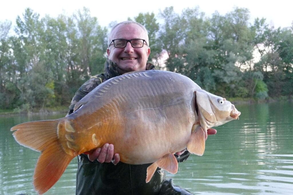 Angler holding large mirror carp by lake.