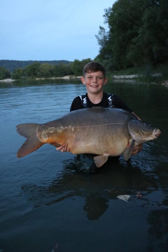 Boy holding large fish in a river at dusk.