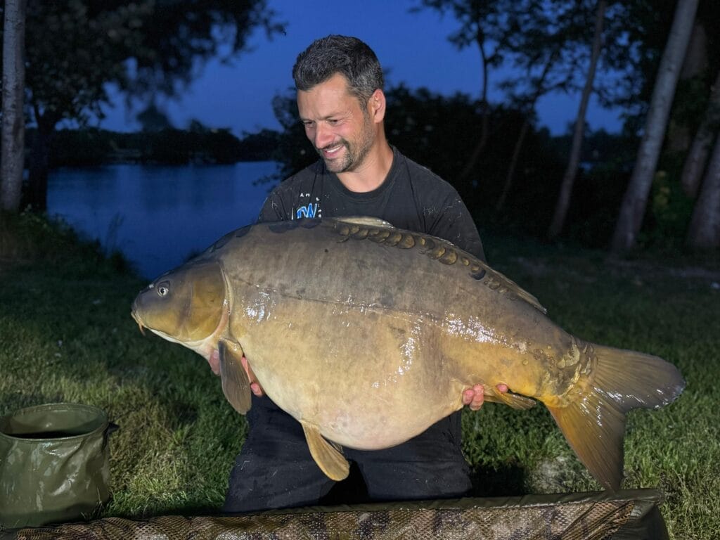 Man holding large carp by a lake.