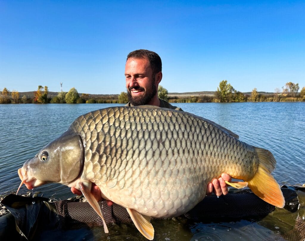 Man holding large carp by a lake.